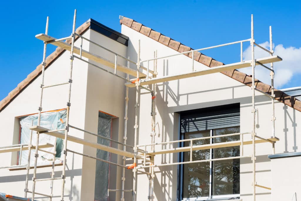 Façade de maison en construction avec échafaudage et crépi clair sous ciel bleu. Fenêtres bâchées et volet fermé, tuiles de toit apparentes.