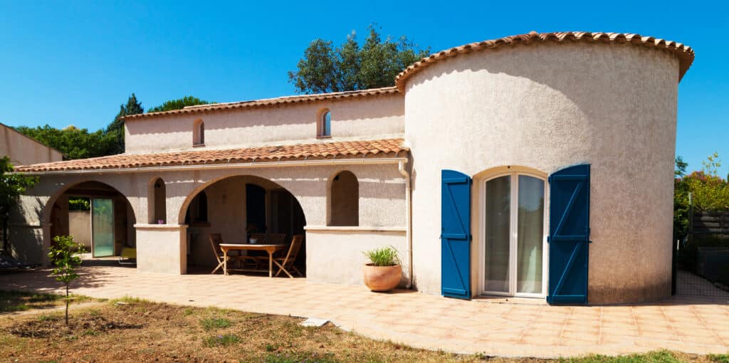 Maison méditerranéenne en crépi clair sous ciel bleu. Volets bleus, toit en tuiles et patio carrelé.