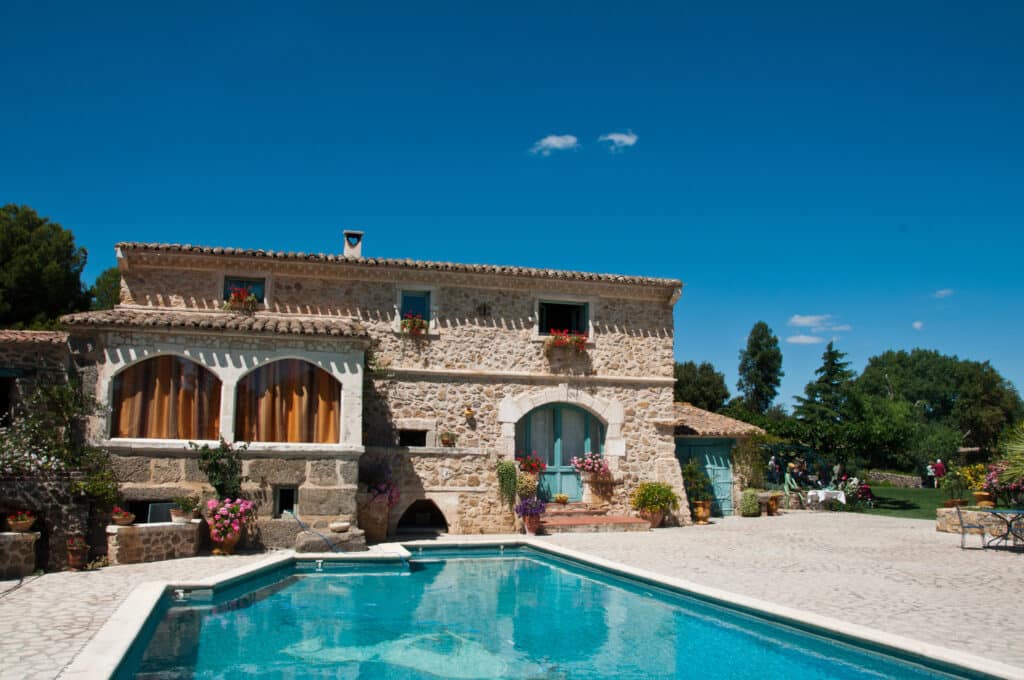 Maison en pierre provençale avec toit en tuiles, cheminée en cœur et fleurs colorées. Grande piscine bleue, terrasse pavée et jardin verdoyant avec invités sous un ciel d'été.