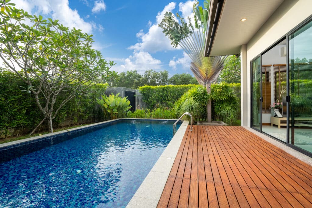 Piscine bleue d'une villa moderne avec terrasse en bois, entourée d'un jardin tropical luxuriant sous un ciel nuageux. Palmier du voyageur distinctif.