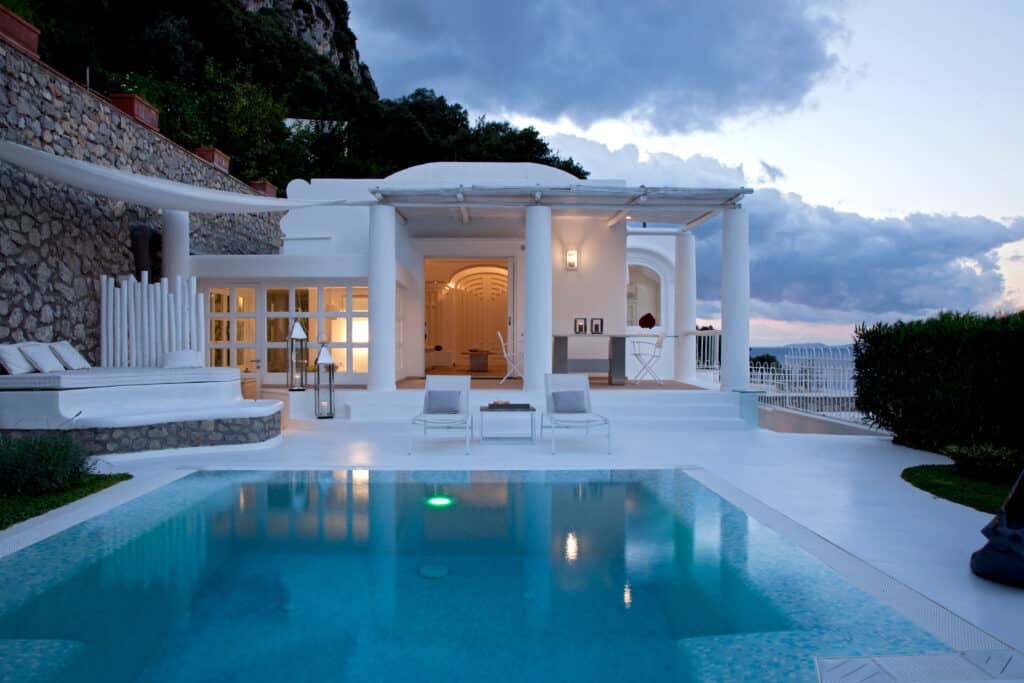 Luxueuse villa blanche méditerranéenne avec piscine illuminée au crépuscule. Terrasse élégante, chaises longues et vue sur la mer et les montagnes.