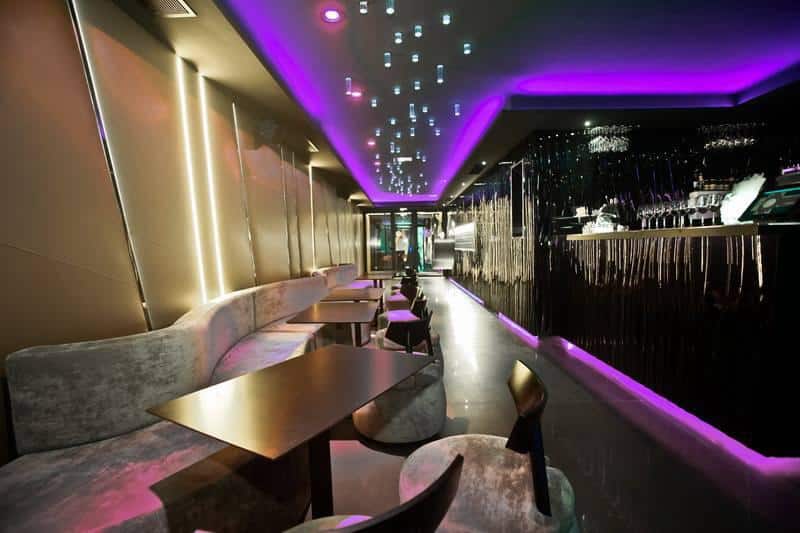 Bar Lounge Moderne Design : Lumières Violettes et Mobilier Chic Intérieur d'un bar lounge moderne avec banquettes grises courbées, tables sombres et éclairage LED violet vif au plafond.