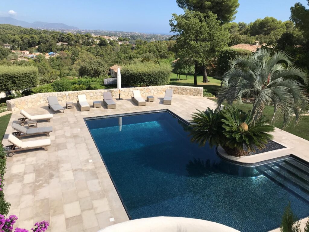 Piscine de luxe avec vue mer et terrasse ensoleillée Piscine à débordement mosaïque bleue sur terrasse carrelée beige, entourée de chaises longues et vue mer panoramique.