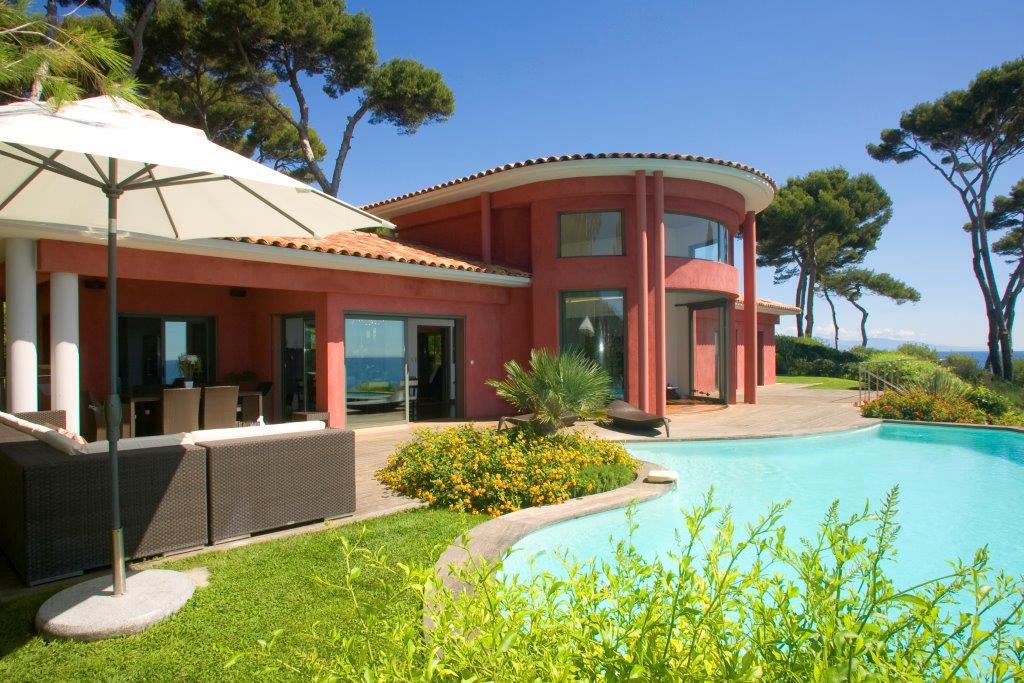Villa Luxe Rouge Vue Mer Piscine Méditerranée Villa rouge avec piscine extérieure, terrasse en bois, mobilier lounge et vue mer sous un ciel bleu, entourée de pins.