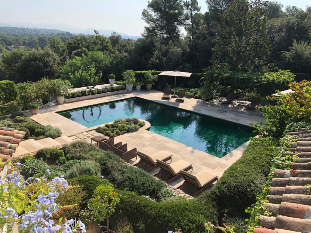 Piscine Luxe Jardin Méditerranéen Vue Panoramique Piscine rectangulaire aux eaux vert émeraude, entourée de dalles de pierre, de chaises longues et d'un jardin méditerranéen luxuriant avec vue sur des collines boisées.