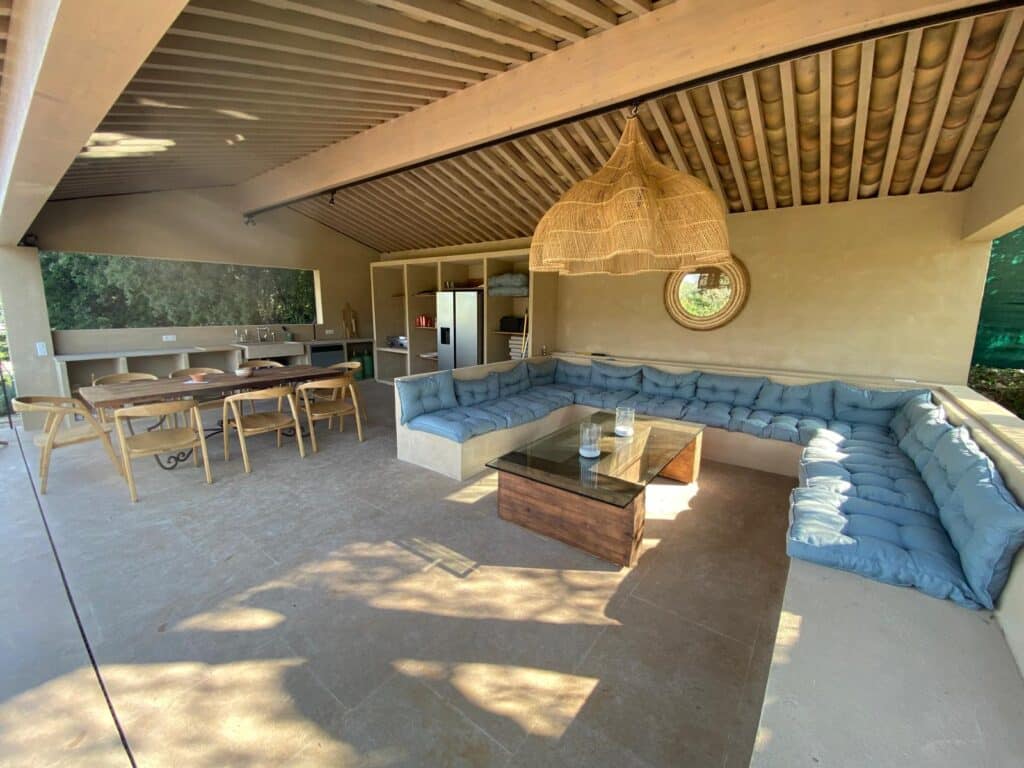 Cuisine d'été moderne avec salon extérieur en L, banquettes bleues et table en bois sous un toit en terre cuite.
