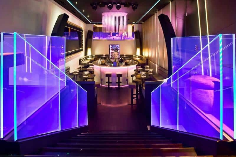 Bar lounge moderne avec éclairage néon violet, bar central circulaire et garde-corps en verre illuminé.
