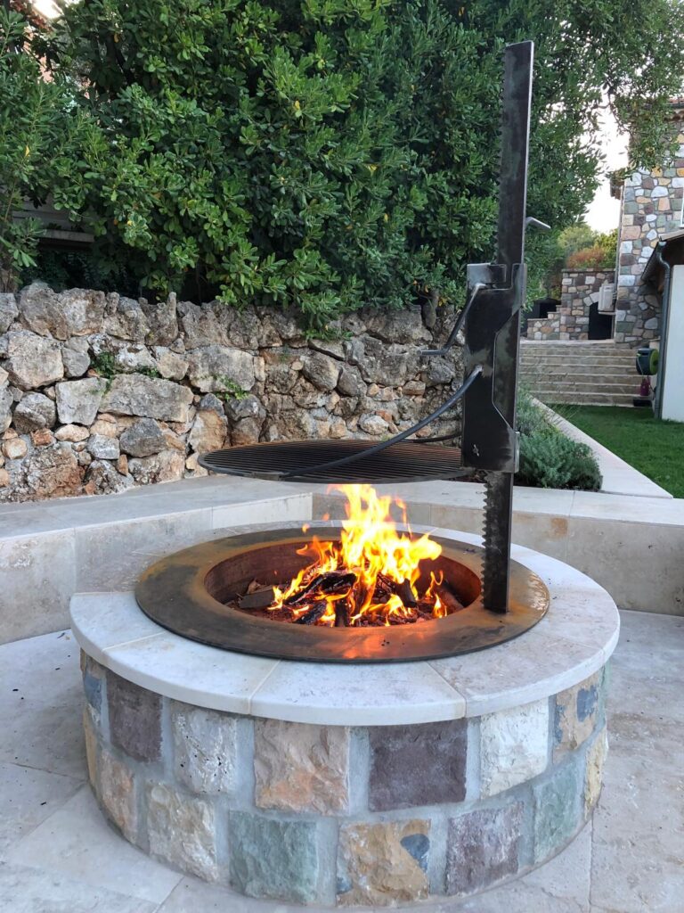 Brasero en pierre naturelle avec feu de bois et grille de barbecue suspendue dans un jardin avec mur de soutènement.