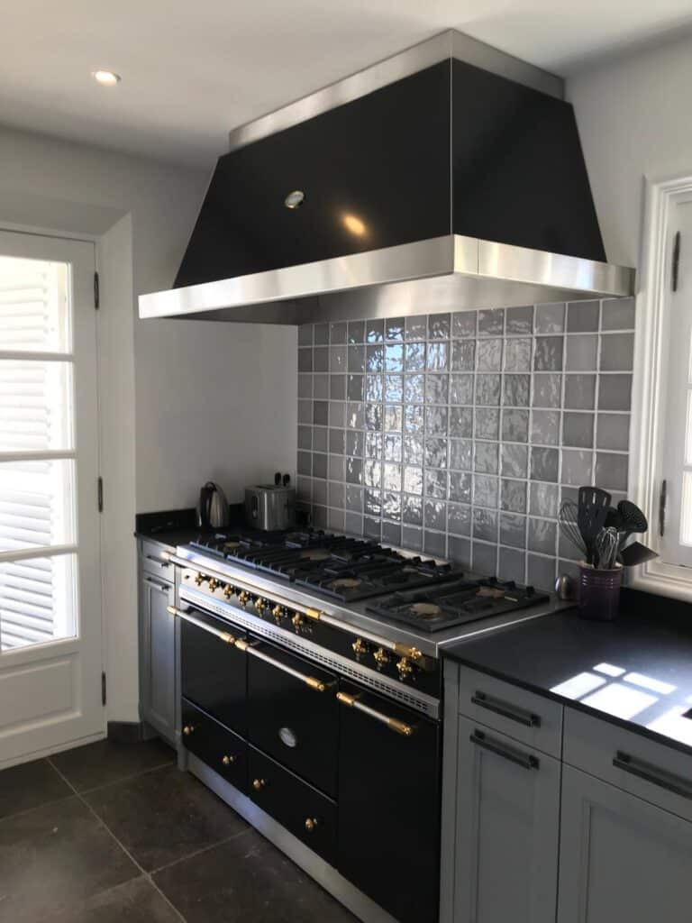 Piano de cuisson Lacanche noir et inox sous une grande hotte, avec crédence en carreaux de zellige gris.