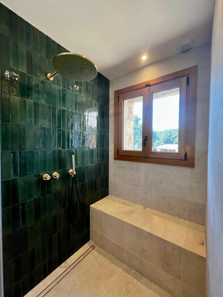 Douche moderne avec carrelage vert foncé, robinetterie en laiton doré, banc en pierre et fenêtre en bois naturel.