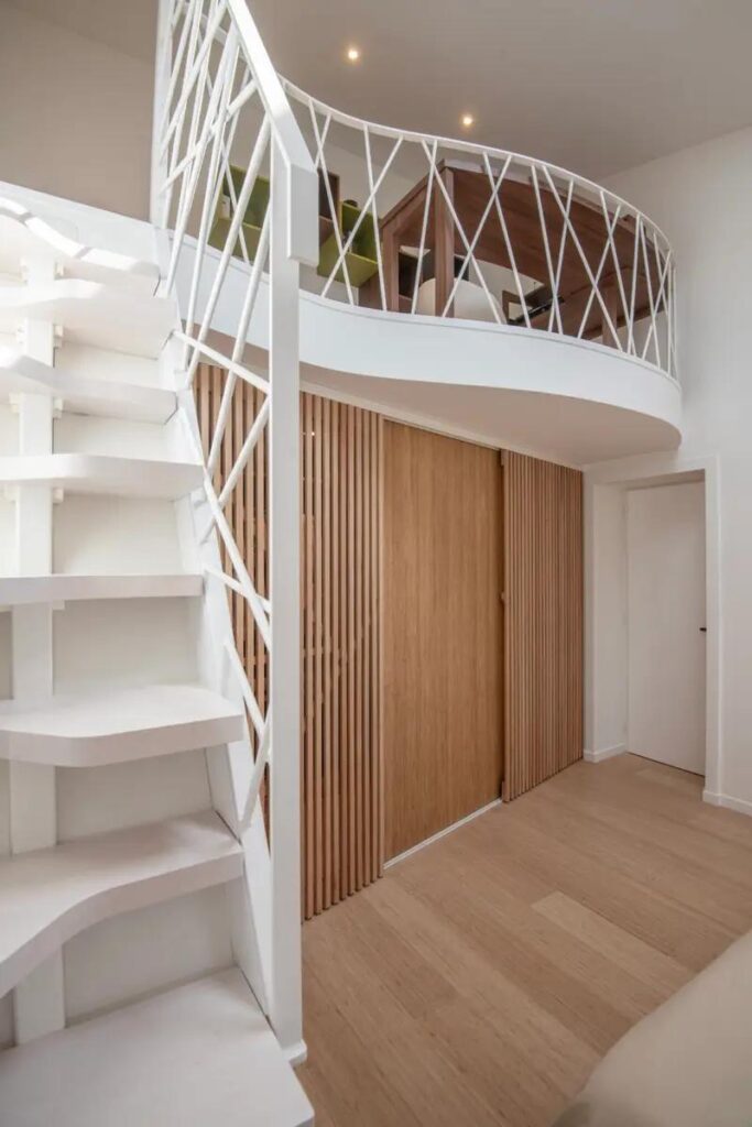 Escalier en colimaçon blanc menant à une mezzanine avec garde-corps géométrique et rangements en bois clair intégrés.