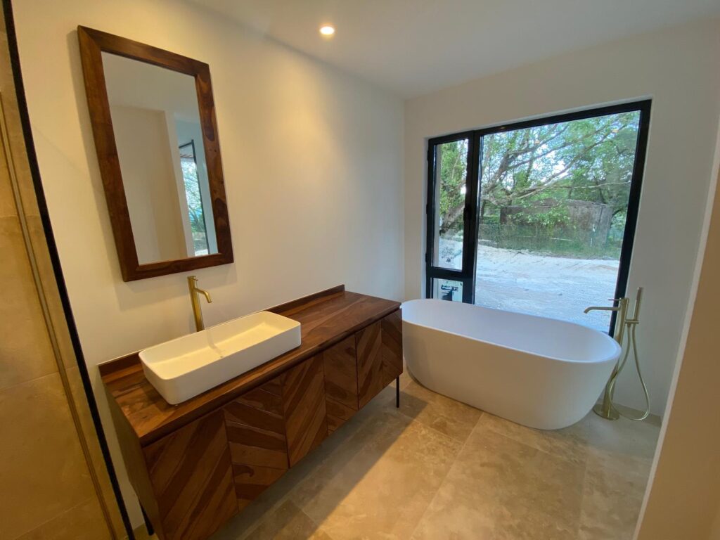 Salle de bain moderne avec meuble en bois, vasque blanche, robinet doré et baignoire îlot devant une grande fenêtre.