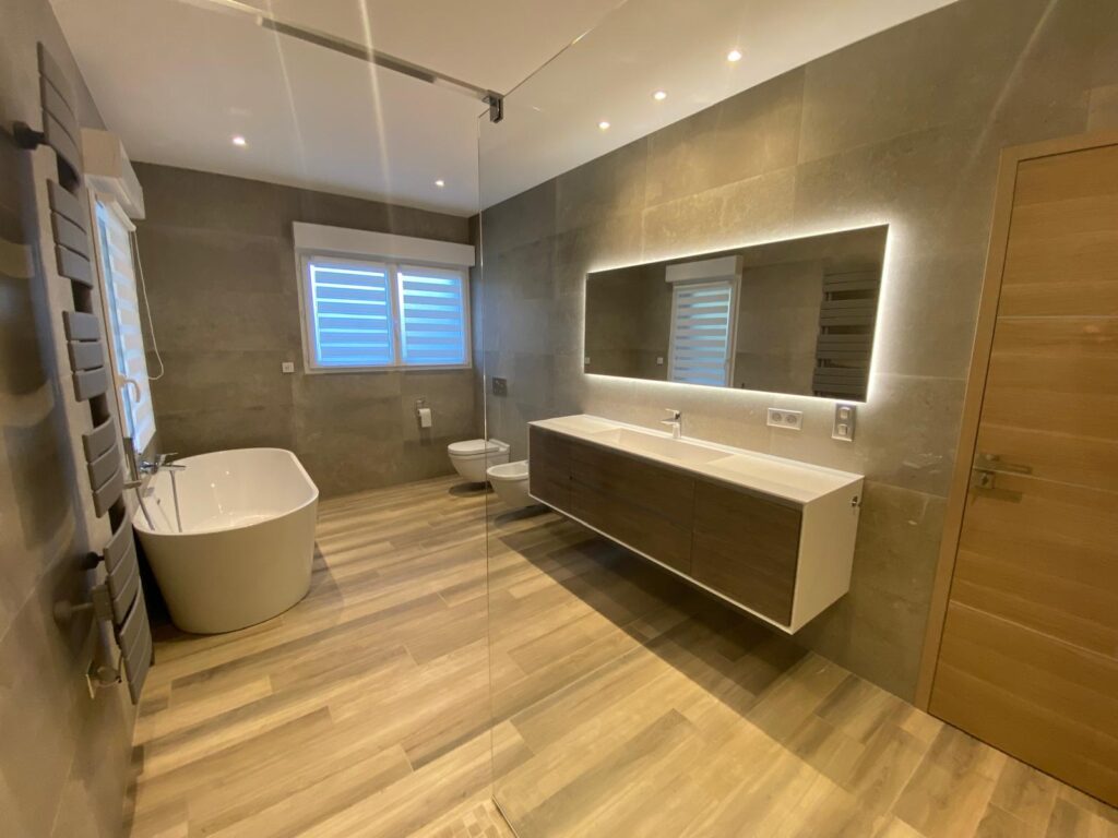 Salle de bain moderne avec baignoire îlot, meuble vasque suspendu, miroir rétroéclairé et carrelage aspect bois.